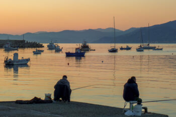 To fiskere med stenger i profil mot kveldsblank sjø - noen småbåter bak